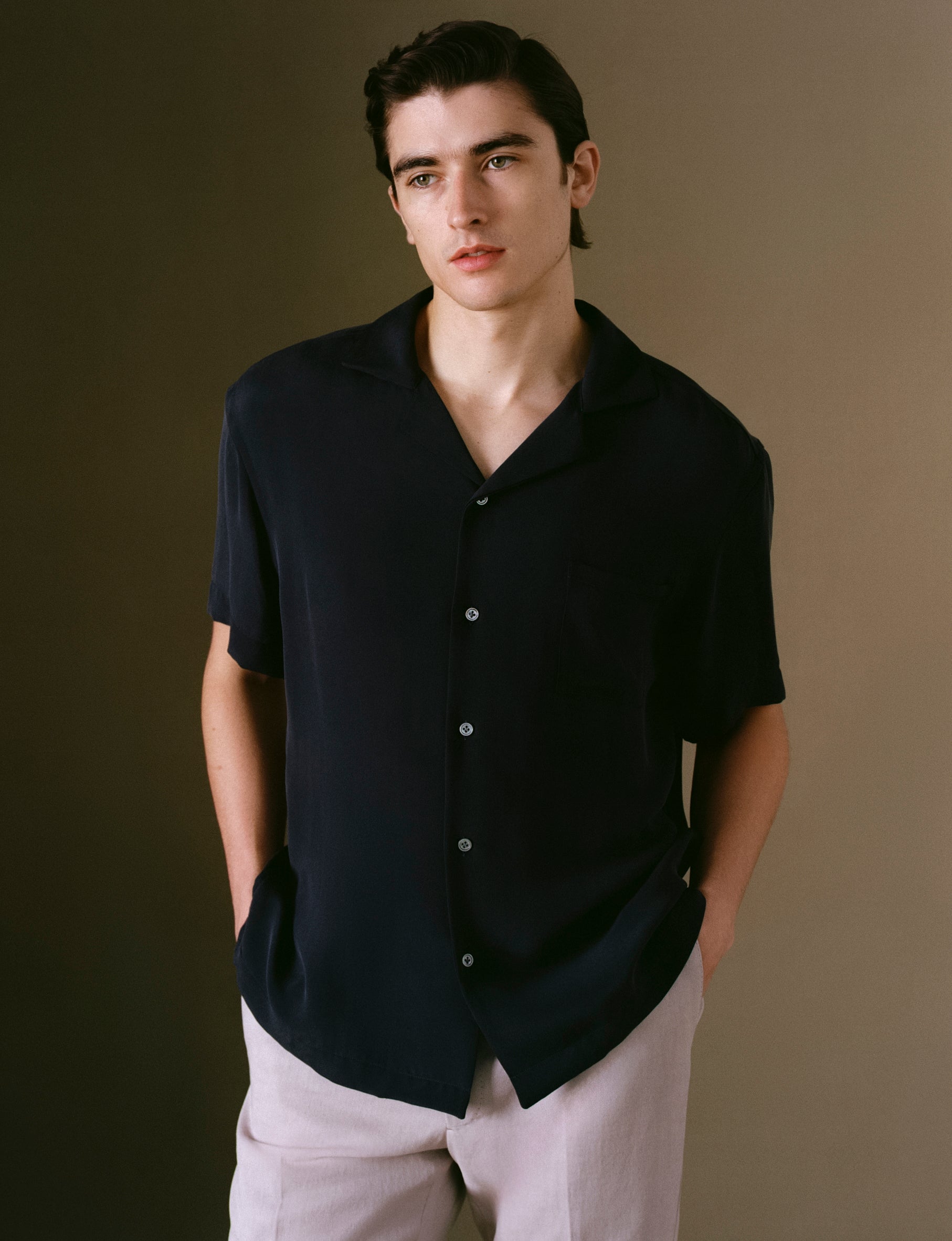 Model wearing a silk navy button-up shirt and light pants against a brown background