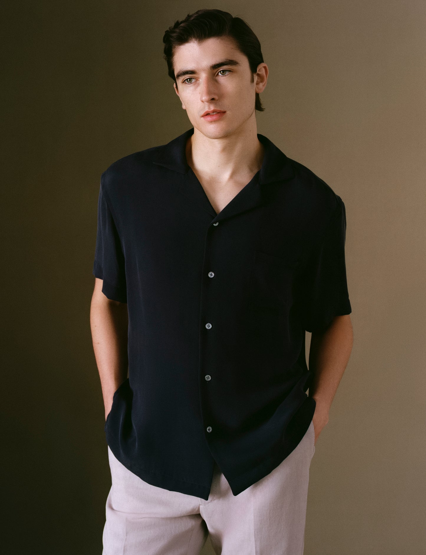 Model wearing a silk navy button-up shirt and light pants against a brown background