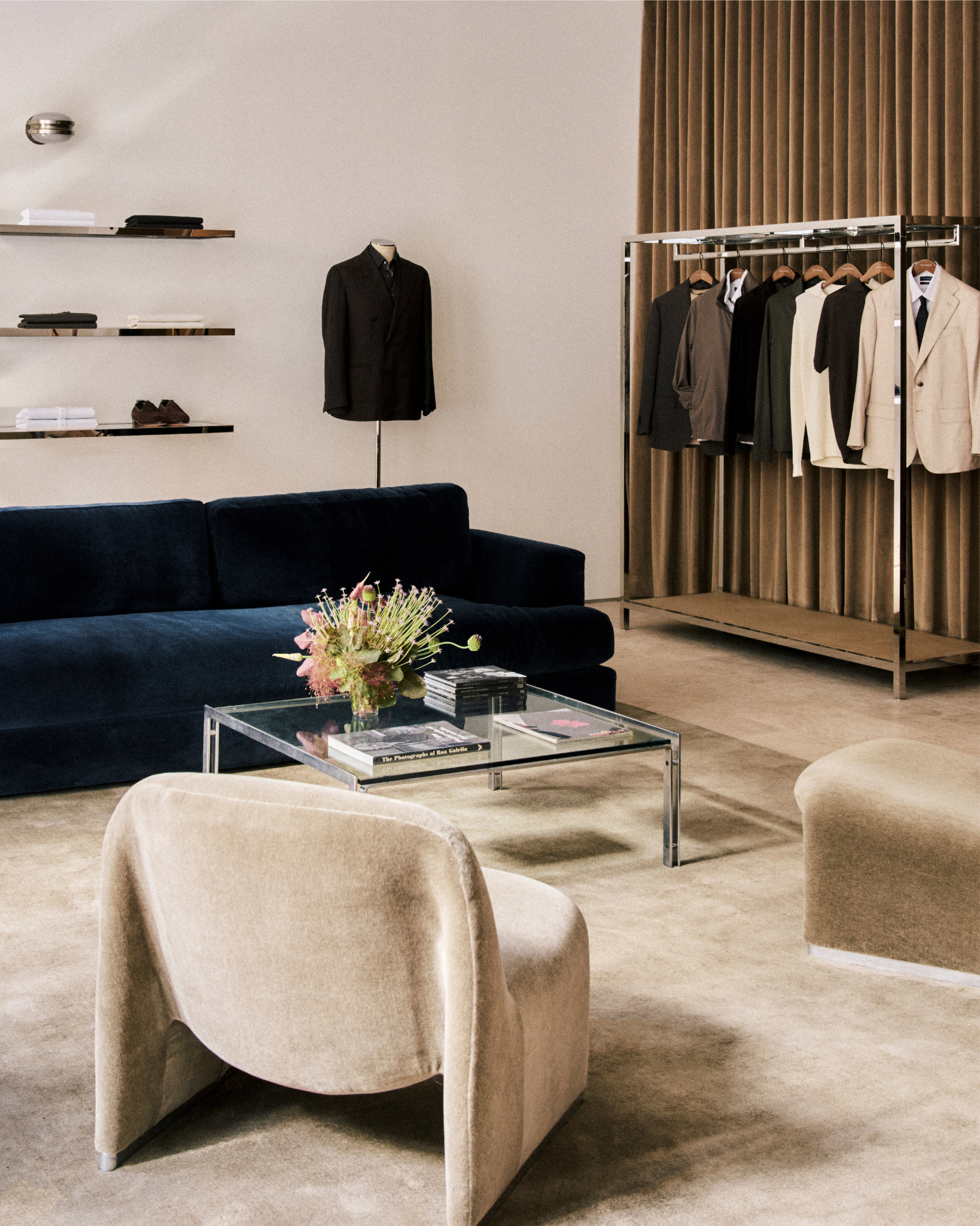 Modern living room with a navy sofa, beige armchair, and a clothing rack with suits.