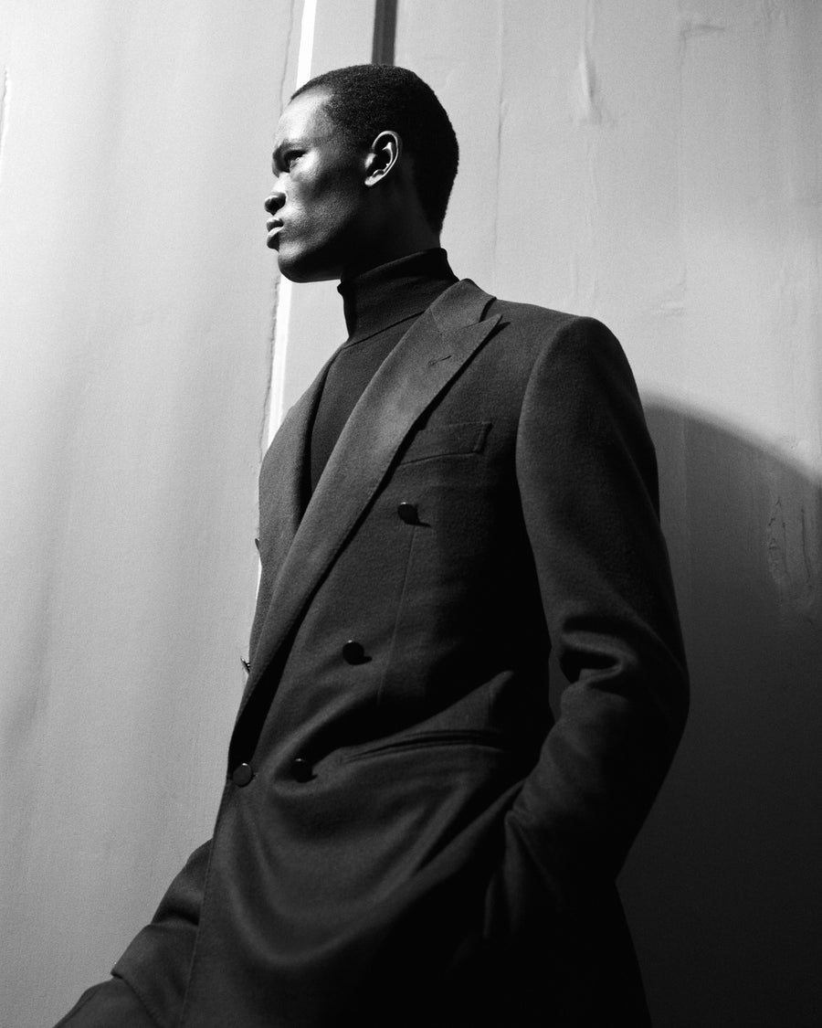 Model wearing a dark double-breasted suit jacket over a black turtleneck, leaning against a textured wall.
