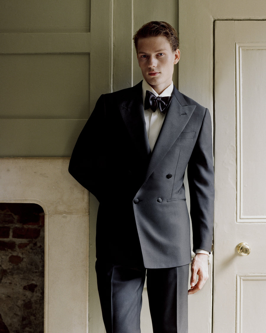 A man wearing a dark double-breasted tuxedo jacket with matching trousers, a white dress shirt, and a black bow tie, standing indoors against a paneled wall and door.