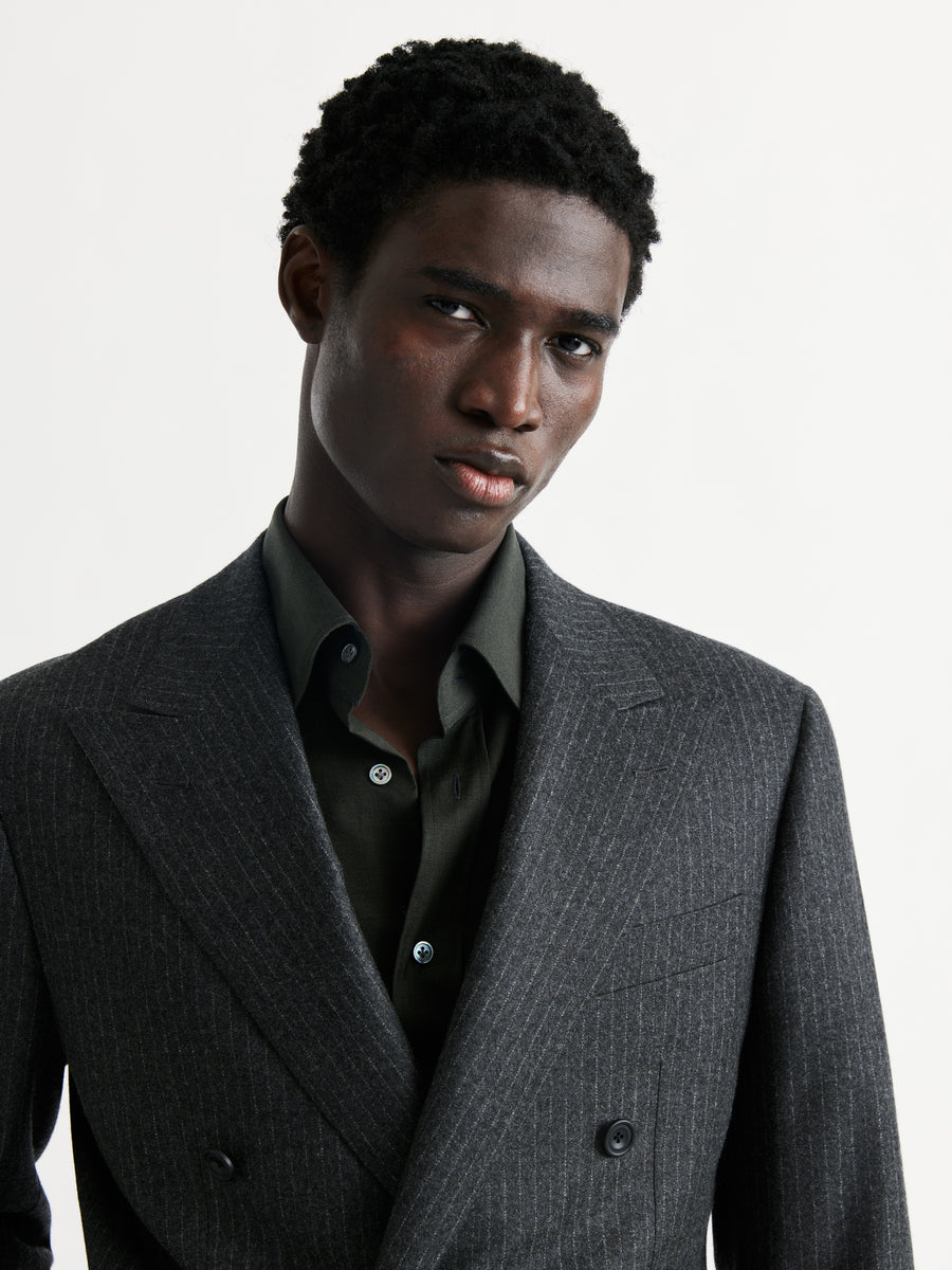 A man wearing a dark gray double-breasted pinstripe suit jacket over a dark olive green button-down shirt, photographed against a clean white studio background.
